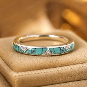Op de afbeelding: Een zilverkleurige bandring met ingelegde segmenten van turkoois steen met zwarte aders, afgewisseld met kleine driehoekige secties die elk drie kleine heldere steentjes bevatten, wat een doorlopend patroon rondom de ring creëert.