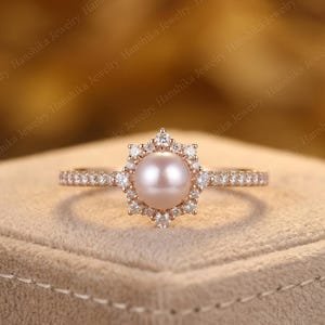 Op de afbeelding: Een roségouden ring met een centrale, lichtroze parel omringd door een halo van kleine, heldere edelstenen. De band is ook bezet met kleine, heldere edelstenen. De ring wordt gepresenteerd tegen een neutrale achtergrond.
