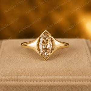May include: A gold ring featuring a marquise-cut champagne-colored gemstone set in a smooth, polished gold band. The setting has a pointed, geometric shape that frames the elongated center stone, creating a sleek and continuous metal profile.