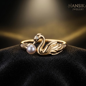 Könnte beinhalten: Ein goldfarbener Ring mit einem Schwan-Design, der eine Perle im Schnabel hält. Der Schwan hat einen kleinen Diamanten. Der Ring ist vor einem dunklen Hintergrund platziert, mit dem Markennamen "HANSIKA JWELLRY".