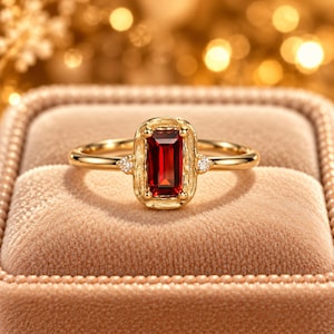 May include: A gold ring featuring a rectangular emerald-cut deep red garnet gemstone set in a textured, organic-style bezel. The band includes a small, round, clear accent stone on each side of the central setting.