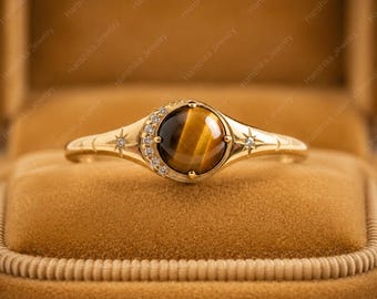 May include: A gold ring featuring a round, polished tiger's eye gemstone at the center. The band is accented with a crescent moon shape set with small clear stones, flanked by engraved star motifs on both sides of the polished gold metal band.
