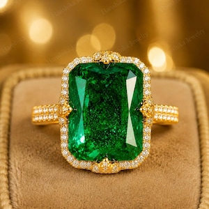 Puede incluir: Un anillo de oro con una gran piedra central rectangular de color verde vibrante rodeada por un halo de pequeñas piedras blancas. La banda también está incrustada con pequeñas piedras blancas y el engaste incluye garras doradas.
