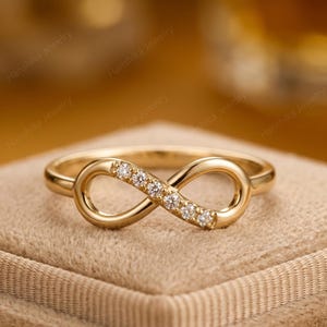 Könnte beinhalten: Goldfarbener Unendlichkeitsring mit einer Reihe kleiner, klarer Edelsteine in der Mitte. Der Ring wird auf einer neutralen Oberfläche mit verschwommenem Hintergrund präsentiert.