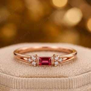 Könnte beinhalten: Roségoldener Ring mit einem rechteckigen Rubin und kleinen Diamantakzenten. Der Ring hat ein zierliches Band und dekorative Diamantcluster auf beiden Seiten des Rubins. Der Ring wird auf einer beigen Oberfläche präsentiert.