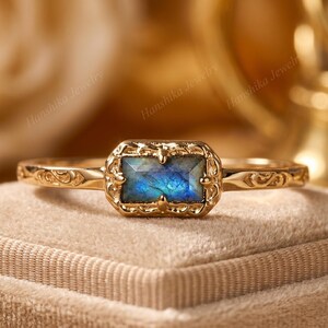 Könnte beinhalten: Ein goldener Ring mit einem rechteckigen, facettierten Labradorit-Edelstein mit blauem Schimmer. Die Fassung hat einen verzierten Metallrand im Filigranstil, und die Ringschiene zeigt aufwendige, gravierte Rankenmuster.