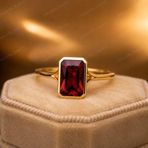 Puede incluir: Un anillo de oro con una gran piedra preciosa granate de color rojo intenso, con talla esmeralda rectangular y engastada en un bisel liso. La banda es fina y pulida, con un diseño de brazo dividido que se une al engaste central.