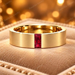 Puede incluir: Un anillo de banda de oro con acabado mate cepillado que presenta una única piedra preciosa roja de corte baguette rectangular engastada verticalmente en el centro. El anillo tiene un perfil plano y un interior pulido de diseño sencillo.