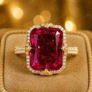 Puede incluir: Un anillo de oro con una gran piedra preciosa rectangular de color rojo intenso en el centro. La piedra está rodeada por un halo de pequeñas piedras claras y brillantes. La banda también está incrustada con piedras claras y es de oro amarillo.