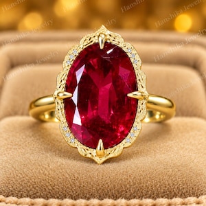 May include: A gold ring featuring a large, oval-cut, deep red gemstone held by four prongs. The setting has an ornate, engraved metal border accented with tiny, clear, round stones. The band is smooth, polished gold, creating a bold, elegant look.