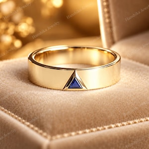 Peut inclure: Une bague anneau en or poli avec une unique petite pierre précieuse bleue de taille triangulaire, sertie à fleur dans le centre de la surface métallique. La bague présente un profil plat et lisse avec une finition réfléchissante.