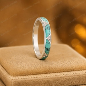 May include: A silver band ring featuring inlaid segments of turquoise stone with black veining, alternating with small triangular sections containing tiny clear stones. The ring has a smooth, polished metallic finish and a slim profile.