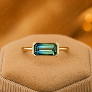 Könnte beinhalten: Ein Goldring mit einem horizontalen Baguette-Schliff-Edelstein, der einen Farbverlauf von Blau- und Grüntönen aufweist. Der Stein ist in einer glatten Goldfassung eingefasst, die die rechteckige Form umrahmt und auf einer schlichten Goldschiene sitzt.