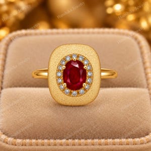 May include: A gold ring featuring a central oval-cut red ruby surrounded by a halo of small round white diamonds. The ruby and diamond setting is encased within a textured, matte-finish gold rectangular frame with rounded corners.