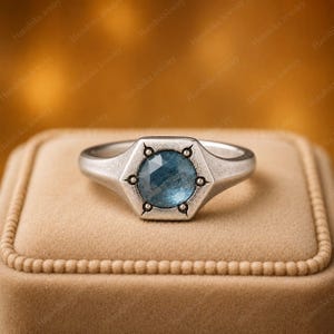 Könnte beinhalten: Ein silberfarbener Ring mit einem runden, facettierten blauen Edelstein in einer sechseckigen Fassung. Die Metallschiene hat eine texturierte, matte Oberfläche und der Stein wird von sechs kleinen, gleichmäßig verteilten Metallkrampen gehalten.