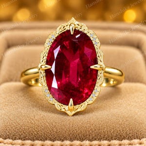 K&ouml;nnte beinhalten: Ein Goldring mit einem gro&szlig;en, oval geschliffenen, tiefroten Edelstein, der von vier Krappen gehalten wird. Die Fassung hat einen verzierten, gravierten Metallrand, der mit winzigen, klaren, runden Steinen besetzt ist. Die Ringschiene ist glatt.