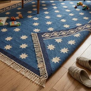May include: A blue area rug featuring a repeating pattern of cream-colored eight-pointed stars. The rug has a dark blue border with a cream diamond geometric design and is finished with long, cream-colored fringe along the edges.