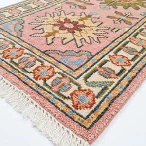 Puede incluir: Una alfombra rosa y crema con un detallado estampado floral. La alfombra presenta un diseño central con grandes flores estilizadas en tonos amarillos, rojos y verdes. El borde incluye motivos florales más pequeños y una franja en un borde.