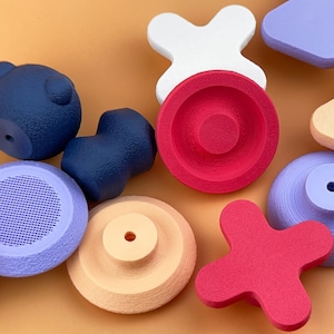 May include: An assortment of colorful, geometric wooden shapes in various colors, including red, blue, white, and tan. The shapes include circles, crosses, and other abstract forms, arranged on a solid orange background. The objects appear to be toys or decorative items.