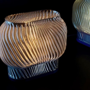 May include: A translucent, oval-shaped lamp with a ribbed, wave-like texture. The structure features a narrow base that widens toward the top, emitting a soft, warm glow from within. The surface is composed of repeating vertical lines.