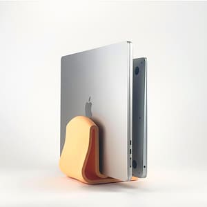 May include: A light orange, curved, U-shaped vertical laptop stand holding two silver laptops in an upright position. The stand features a smooth, matte finish and a wide base that provides stability for the electronic devices stored within its slot.