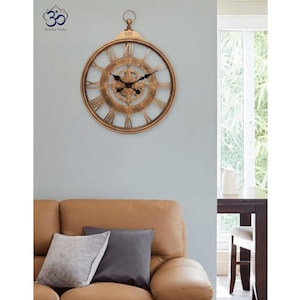 May include: A round wall clock with a metallic gold finish, featuring a compass-inspired center design, black hands, and large Roman numerals. The clock has a circular hanging loop at the top and includes the text Divine Veda with an Om symbol.
