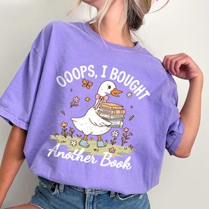 May include: A violet-colored t-shirt with the text "OOOPS, I BOUGHT Another Book." The graphic features a white duck carrying books, wearing a pink bow, and blue boots, surrounded by flowers and butterflies.