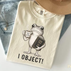May include: Ivory-colored t-shirt featuring a frog holding a coffee cup and a book labeled "CASE FILE." The text "I OBJECT!" is printed below the frog. The shirt is laid on top of blue jeans and a tan hat.