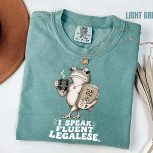 Pode incluir: Camiseta verde claro com um desenho de um sapo segurando uma xícara de café e um livro com a legenda "Case File". O texto "I Speak Fluent Legalese" está impresso abaixo. Um colar de prata está sobre a camiseta.