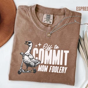 May include: Espresso-coloured t-shirt featuring a goose in sunglasses on a scooter, with the text "Off to Commit Mom Foolery". A silver necklace with a pendant is draped on the shirt. The shirt is a size S-CH.