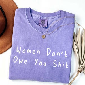 Puede incluir: Una camiseta de color lila con el texto blanco "Women Don't Owe You Shit". Un collar dorado con un colgante está sobre la camiseta. Un sombrero marrón y una hoja de palma seca están en el fondo.