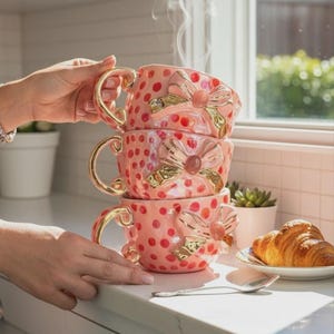 Puede incluir: Una pila de tres tazas de té rosas de cerámica con asas doradas y detalles de lazo. Cada taza está decorada con lunares rojos. Sale vapor de la taza superior. Un cruasán está en un plato cercano.