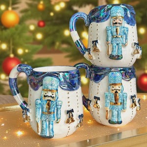May include: Three white ceramic mugs with blue and iridescent accents. Each mug features a blue nutcracker soldier design and gold bell embellishments. The mugs are stacked and arranged on a gold surface.