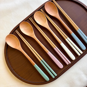 May include: A wooden serving tray holds six wooden spoons and six pairs of chopsticks. The chopsticks have painted tips in pastel colors: blue, white, pink, and green. The spoons and tray are a warm brown.