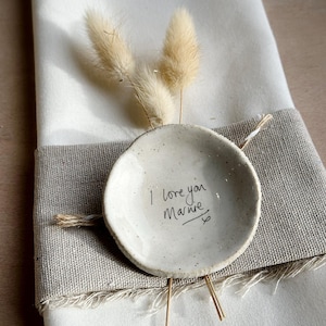 Puede incluir: Un pequeño plato de cerámica moteada con el mensaje escrito a mano "I love you Marnie" y un corazón. El plato está adornado con hierba de cola de conejo seca y descansa sobre una servilleta de lino con un borde de arpillera.