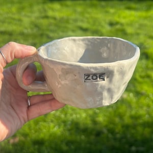 Op de afbeelding: Een handgemaakte, gebroken witte keramische mok met een getextureerd oppervlak en een handvat. Het woord "ZOE" is op de zijkant van de mok gestempeld. De mok wordt tegen een achtergrond van groen gras gehouden.