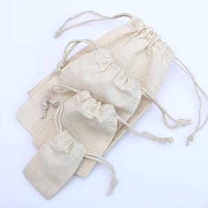 Puede incluir: Un conjunto de cuatro bolsas con cordón de color blanco roto hechas de tela tejida. Cada bolsa presenta un cierre fruncido en la parte superior con cordones trenzados a juego en ambos lados. Las bolsas están dispuestas en una pila escalonada.