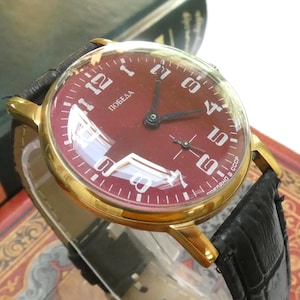 Könnte beinhalten: Eine goldfarbene Armbanduhr mit tiefrotem Zifferblatt, weißen arabischen Ziffern und schwarzen Zeigern. Das Zifferblatt zeigt den kyrillischen Schriftzug POBEDA sowie den Text CDEJLAHO B CCCP. Sie hat ein schwarzes Lederarmband mit Struktur.