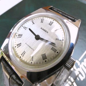 Könnte beinhalten: Eine silberfarbene Karex-Armbanduhr mit rundem Zifferblatt und schwarzen Zeigern. Die Uhr hat römische Ziffern als Stundenmarkierungen und das Wort "ANTIMAGNETIC" auf dem Zifferblatt. Sie hat ein schwarzes Lederarmband.