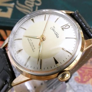 Op de afbeelding: Een goudkleurig polshorloge met een ronde crèmekleurige wijzerplaat, voorzien van de tekst ZentRa, 17 RUBIS en BRUCHSICHER. Het horloge heeft dunne gouden wijzers, een cijfer 12, streepindexen en een zwarte lederen band met textuur.