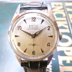May include: A vintage Estia wristwatch with a silver-toned dial, featuring gold-colored Arabic numerals at the 12, 10, 8, 6, 4, and 2 positions. The watch face displays the text Estia, 15 Jewels, and Antimagnetic, with a black leather strap.