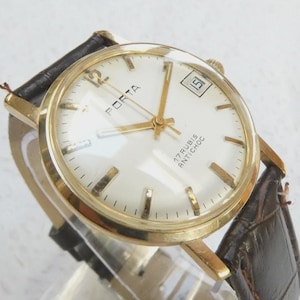 May include: A vintage gold wristwatch with a white face, displaying the brand name "PORTA" and a date window. The watch has gold hands and markers, a leather strap, and the text "17 RUBIS ANTICHOC".