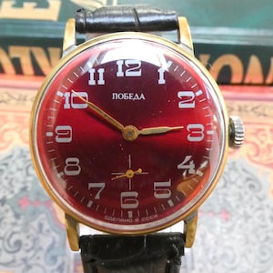 May include: Vintage wristwatch with a burgundy face, gold-tone case, and black leather band. The watch face features white numerals and the word "Победа". The watch has a small seconds dial and the text "СДЕЛАНО В СССР".