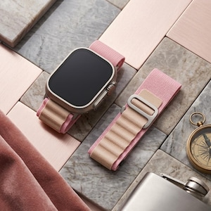 May include: A smartwatch with a black screen and a light pink and tan band is displayed next to a matching band. The watch and band are on a surface with pink and gray tiles. A compass and flask are also visible.