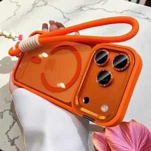 May include: Orange phone case with a matching orange strap. The case has a clear back with an orange border and a camera lens protector. The strap is made of a thick, woven material.