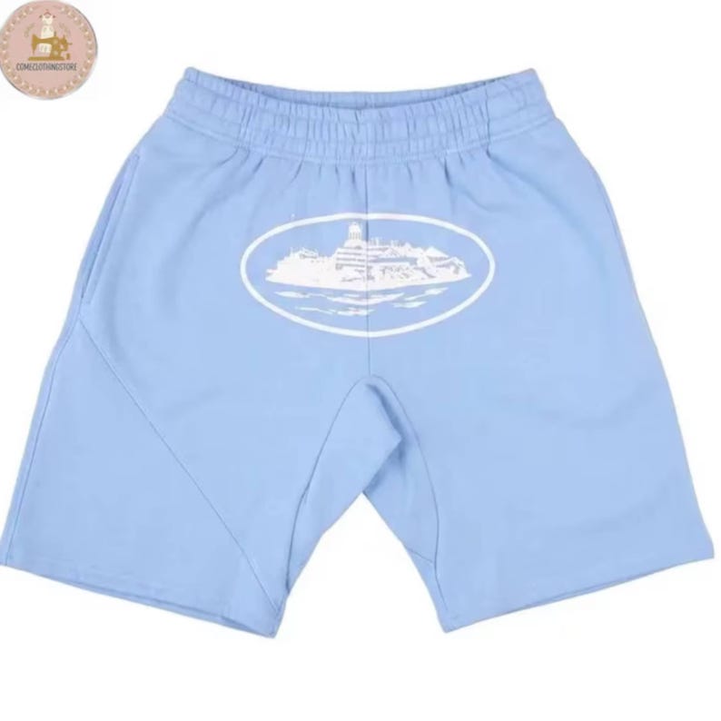 May include: Light blue shorts featuring an elastic waistband and a white oval graphic on the front. The graphic depicts a detailed illustration of a building complex situated on an island surrounded by water, rendered in a clean white line art style.