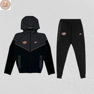 以下が含まれることがあります： 黒色のナイキトラックスーツセット。ジャケットにはフードとジッパーが付いており、胸にはゴールドカラーのロゴ、パンツにはお揃いのロゴが付いています。トラックスーツは柔らかく快適な素材で作られています。