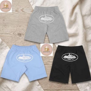 May include: Three pairs of shorts in gray, light blue, and black, each featuring an elastic waistband and a white oval graphic on the front depicting a cruise ship sailing on water. The shorts are arranged in a triangular formation on a fabric surface.