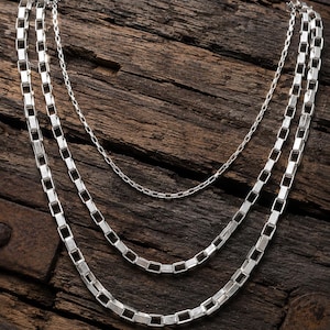 Puede incluir: Un collar de plata de varias hebras que presenta dos cadenas exteriores con eslabones rectangulares y una cadena interior con eslabones cuadrados más pequeños y delicados. El acabado plateado metálico refleja la luz.