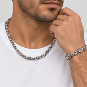 Op de afbeelding: Een zilverkleurige metalen ketting en een bijpassende armband, beide voorzien van een reeks in elkaar grijpende ovale schakels met een gepolijste afwerking. De sieradenset wordt gedragen over een effen wit t-shirt en toont het reflecterende oppervlak.